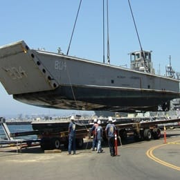 Military landing craft being lifted by crane onto a flatbed trailer at a dock, with crew members supervising the operation